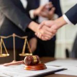 Two people shaking hands over a contract with a gavel and scales nearby.