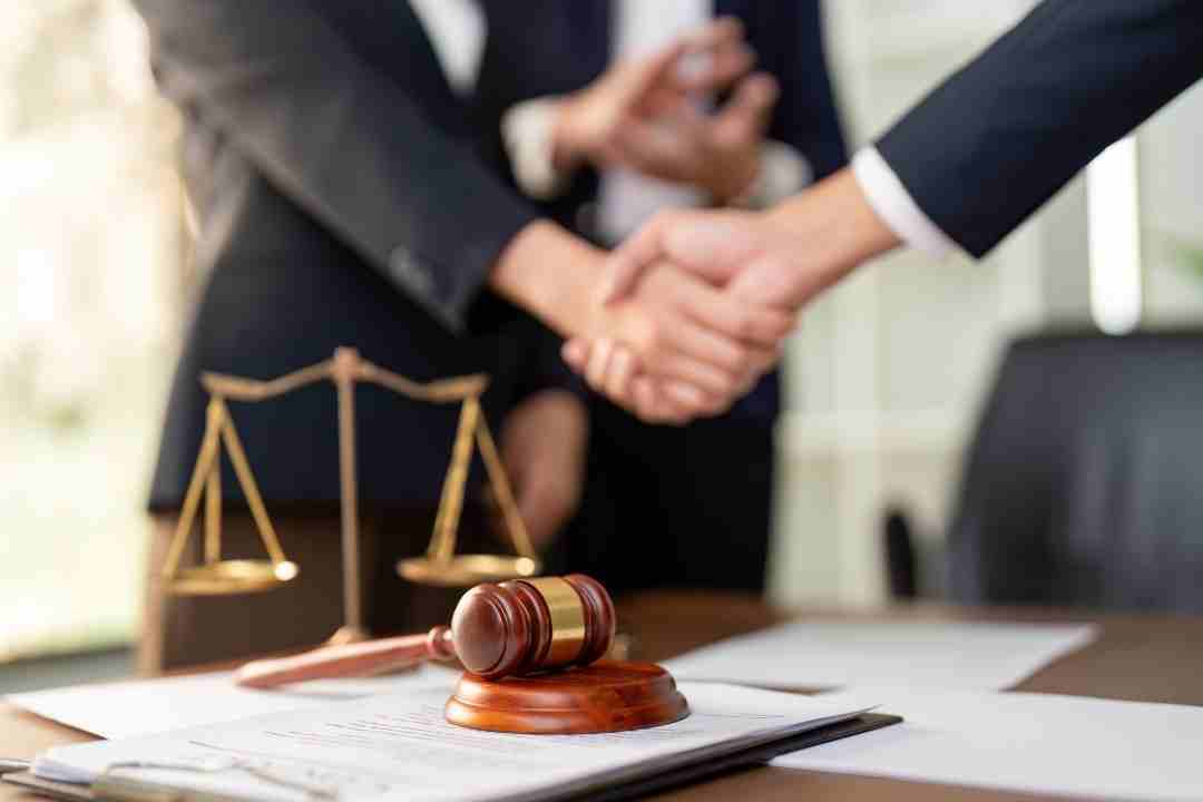 Two people shaking hands over a contract with a gavel and scales nearby.